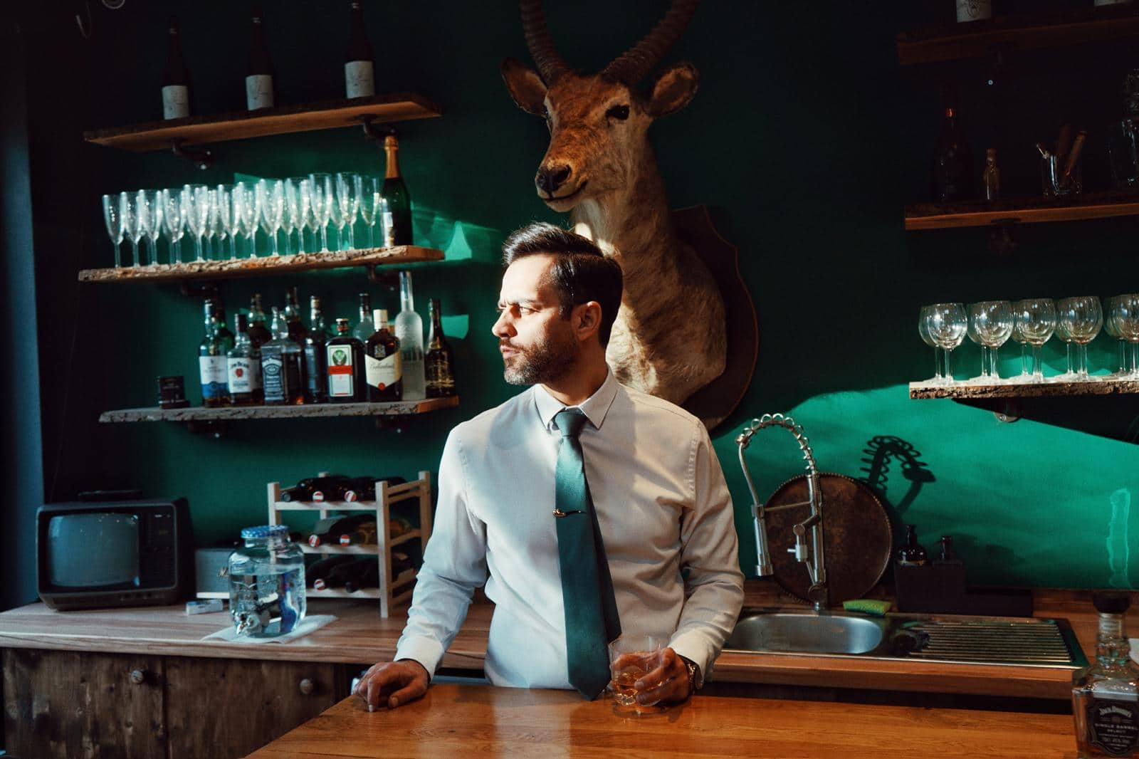 Shouka hinter der Bar seines Barbershops in Wien, vor dunkelgr&uuml;ner Wand mit Wasserbock-Troph&auml;e